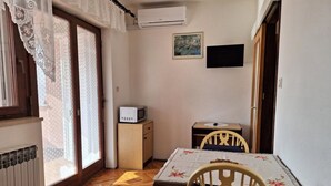 A room with a wooden floor, a dining table with chairs, a small cabinet with a microwave, and a window with lace curtains. - Apartman Štefica with AC and balcony for 2 people (Punat)