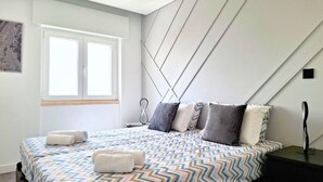 A modern bedroom with a large bed, geometric-patterned bedding, a bedside table, and a window with blinds. - PRAIA DA ROCHA - BELO HORIZONTE (Portimão)