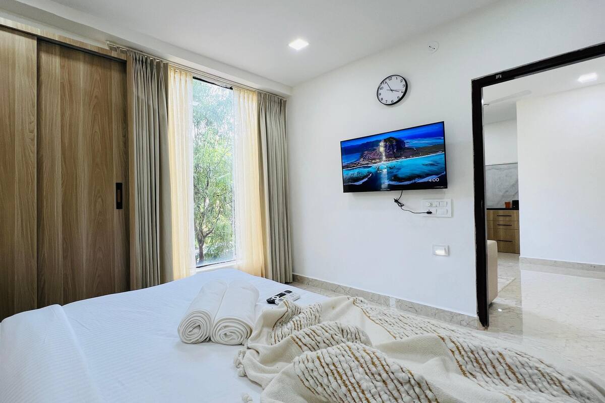 A hotel room with a bed, a TV on the wall, a clock, and a view of the outdoors.