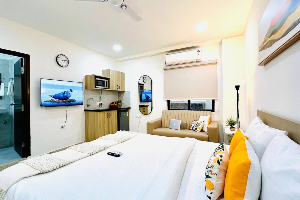 A modern hotel room with a bed, bedside table, TV, and a small kitchenette.