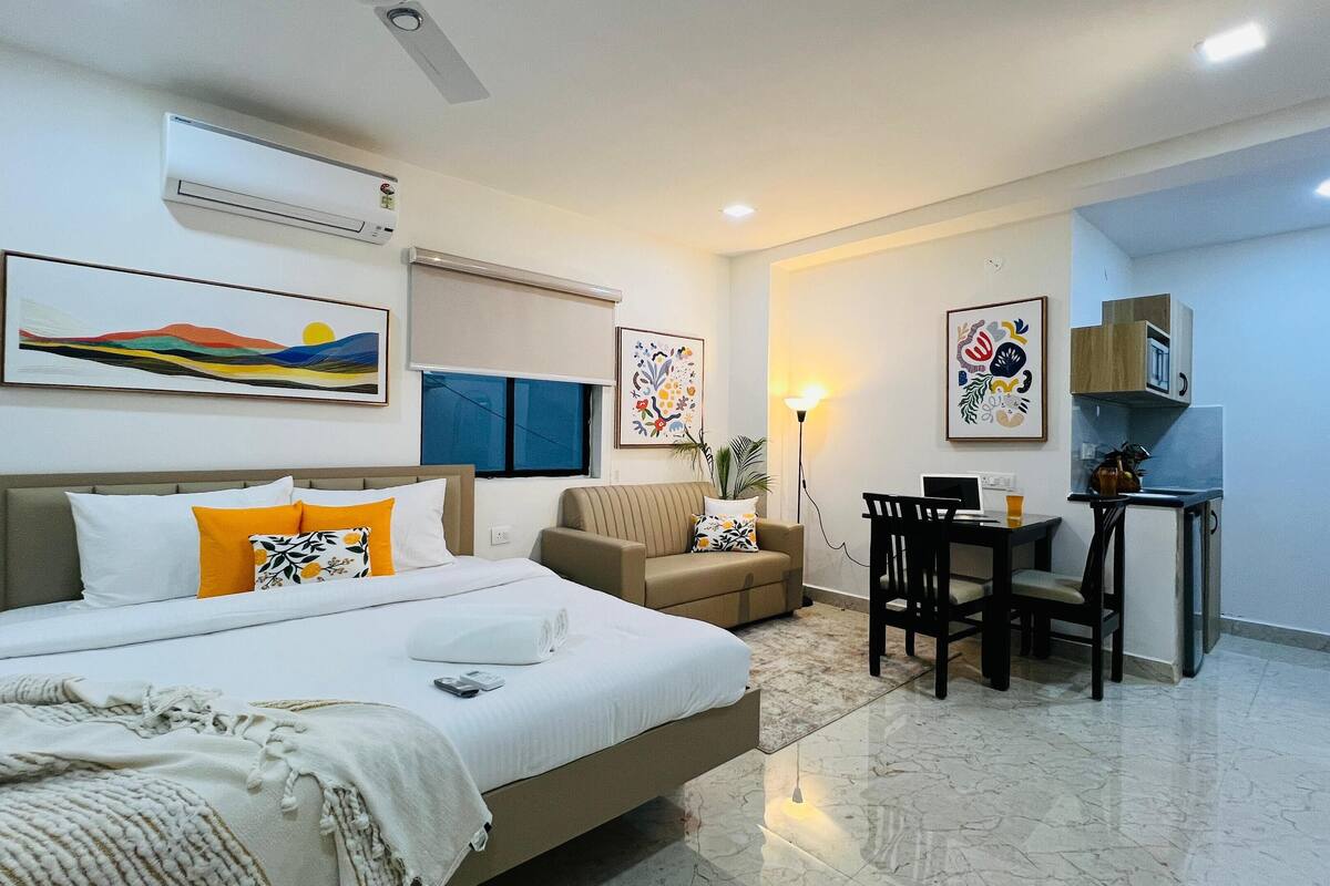A modern hotel room with a bed, a sofa, a desk, and a chair. There is a window, a wall-mounted air conditioner, and artwork on the walls.