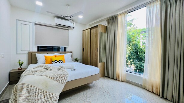 A bedroom with a bed, a ceiling fan, an air conditioning unit, a window with curtains, and a view of greenery outside. - Olive Service Apartments - HICC Hitex Hyderabad (Hyderabad)