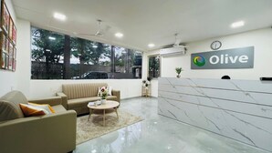 A modern reception area with a marble reception desk, a sofa, a coffee table, and a large window with a view of trees and a building. - Olive Service Apartments - HICC Hitex Hyderabad (Hyderabad)