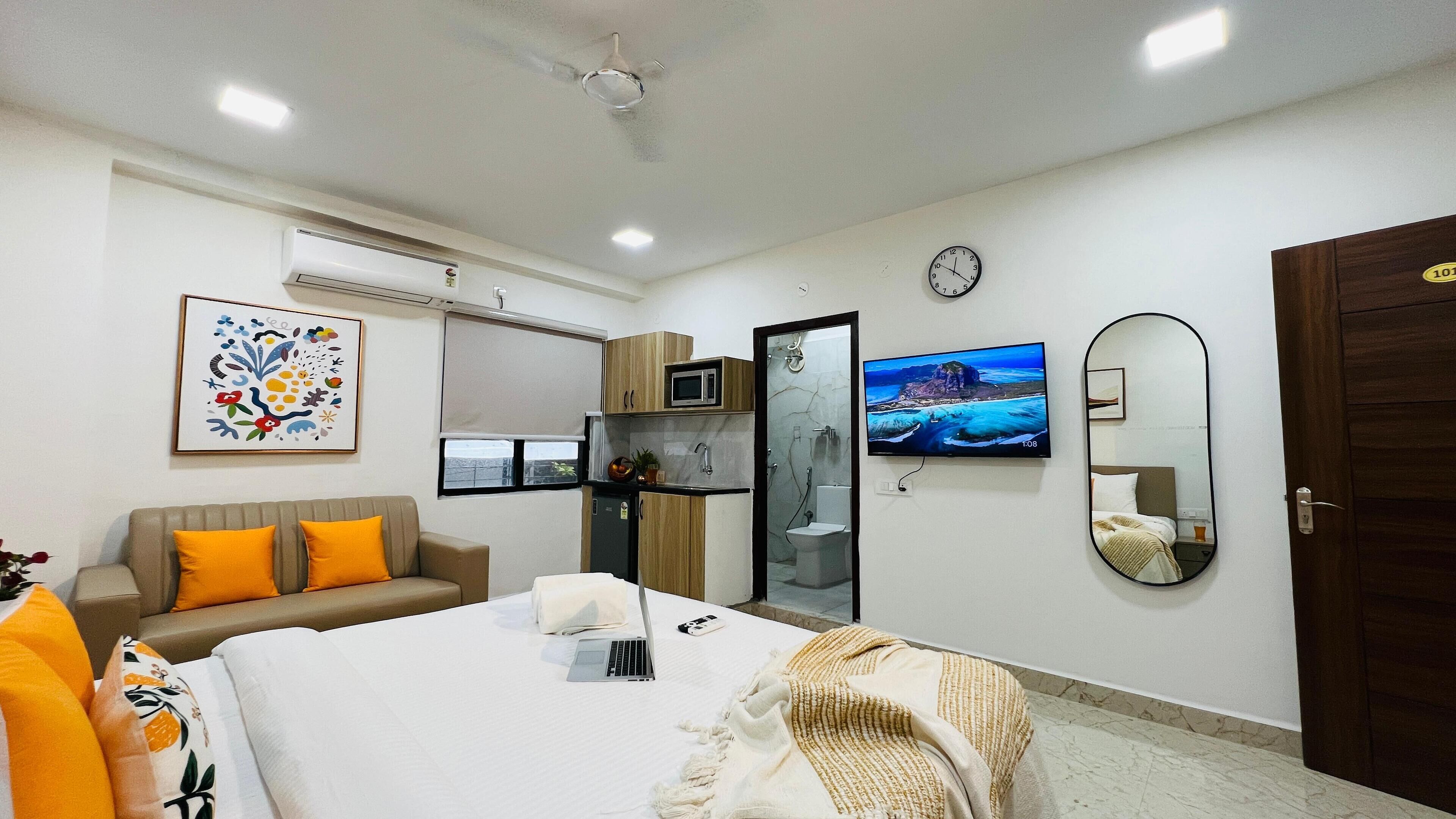A hotel room with a bed, a sofa, a TV, a mirror, a microwave, and a bathroom visible through a glass door.