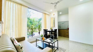 A modern living room with a sofa, a glass coffee table, and a dining area with a table and chairs. There is a ceiling fan, a window with curtains, and a view of greenery outside. - Olive Service Apartments - HICC Hitex Hyderabad (Hyderabad)