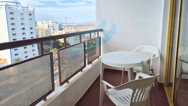 Uma varanda com mesa e cadeiras brancas, guarda-corpo de vidro e vista para edifícios e uma grua de construção.