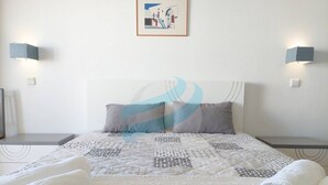 Cama bem arrumada com colcha estampada, dois travesseiros e toalhas dobradas em uma parede branca, com um quadro emoldurado acima.