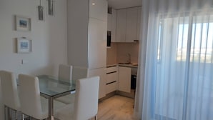 Eine moderne Küche mit weißen Schränken, einem Glastisch und Stühlen. Ein Fenster mit durchsichtigen Vorhängen.