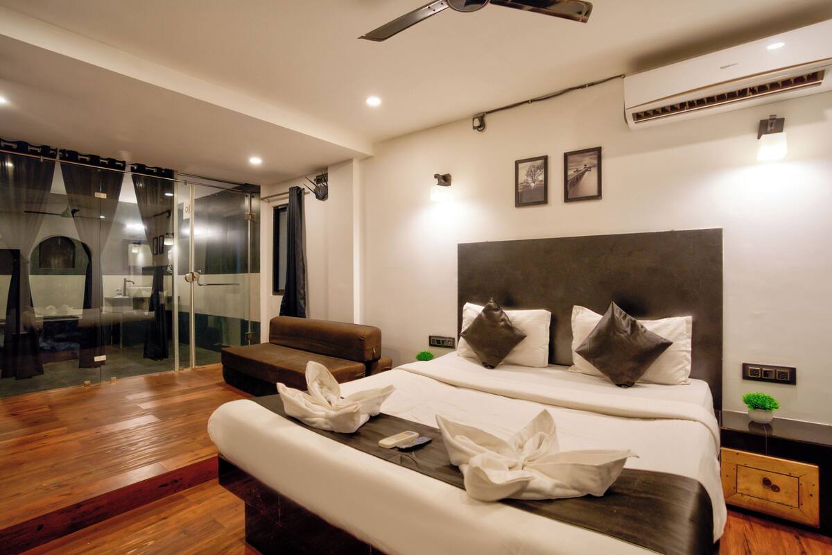 A modern hotel room with a large bed, a separate seating area, a glass shower enclosure, and a ceiling fan.