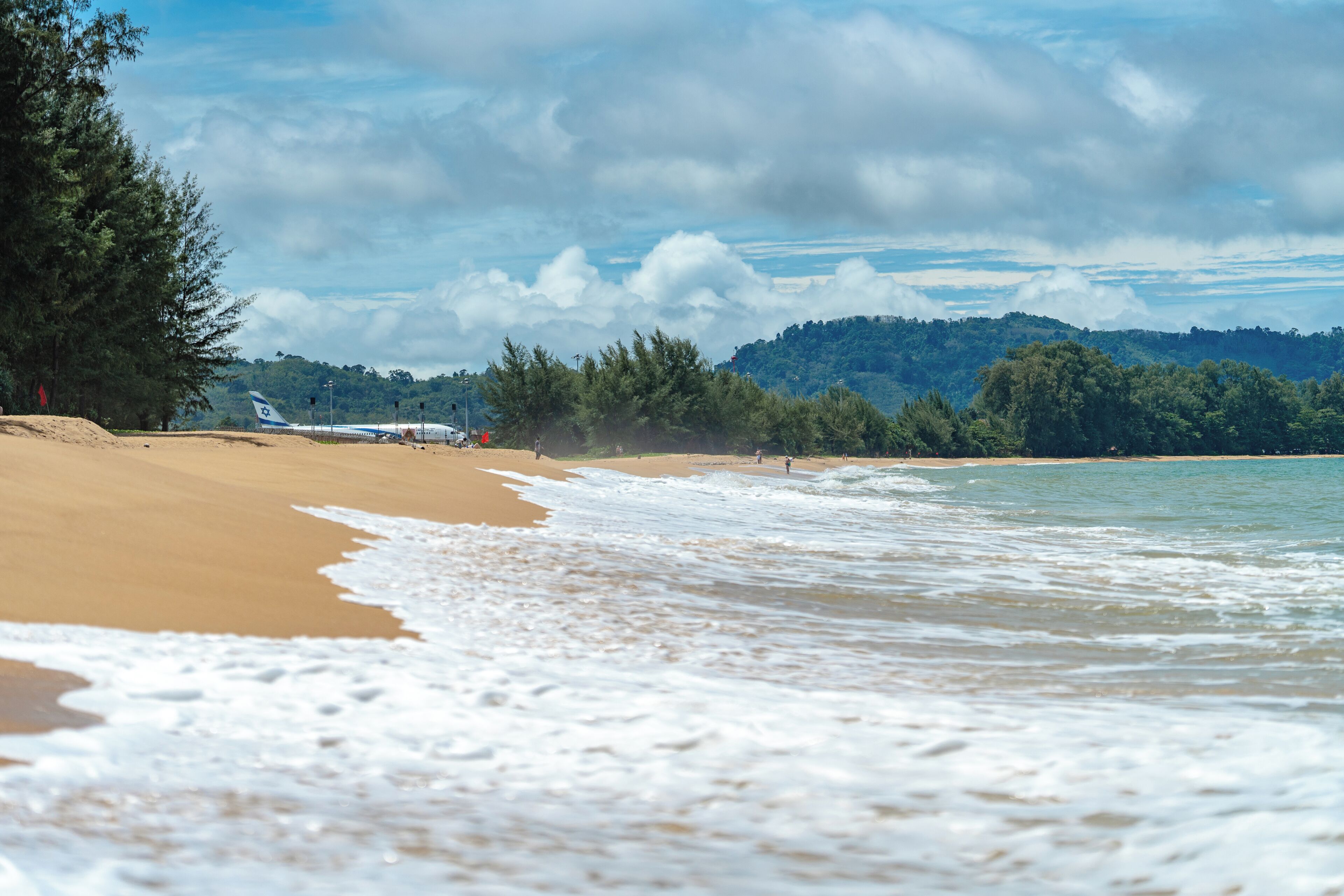 A beach with sand, waves, and a forested hill in the background.