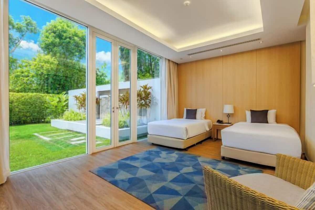A modern hotel room with two beds, a large sliding glass door to a garden, and a wicker chair.