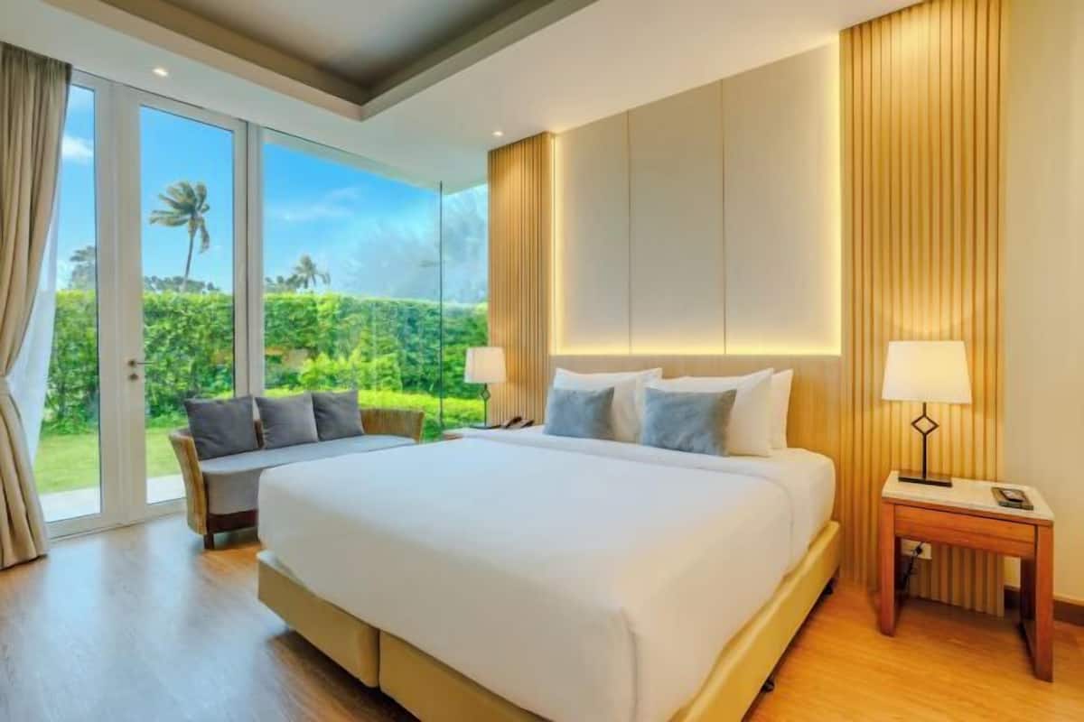 A large bed with a headboard, a bedside table with a lamp, a chair, and a view of a lush garden through large windows.