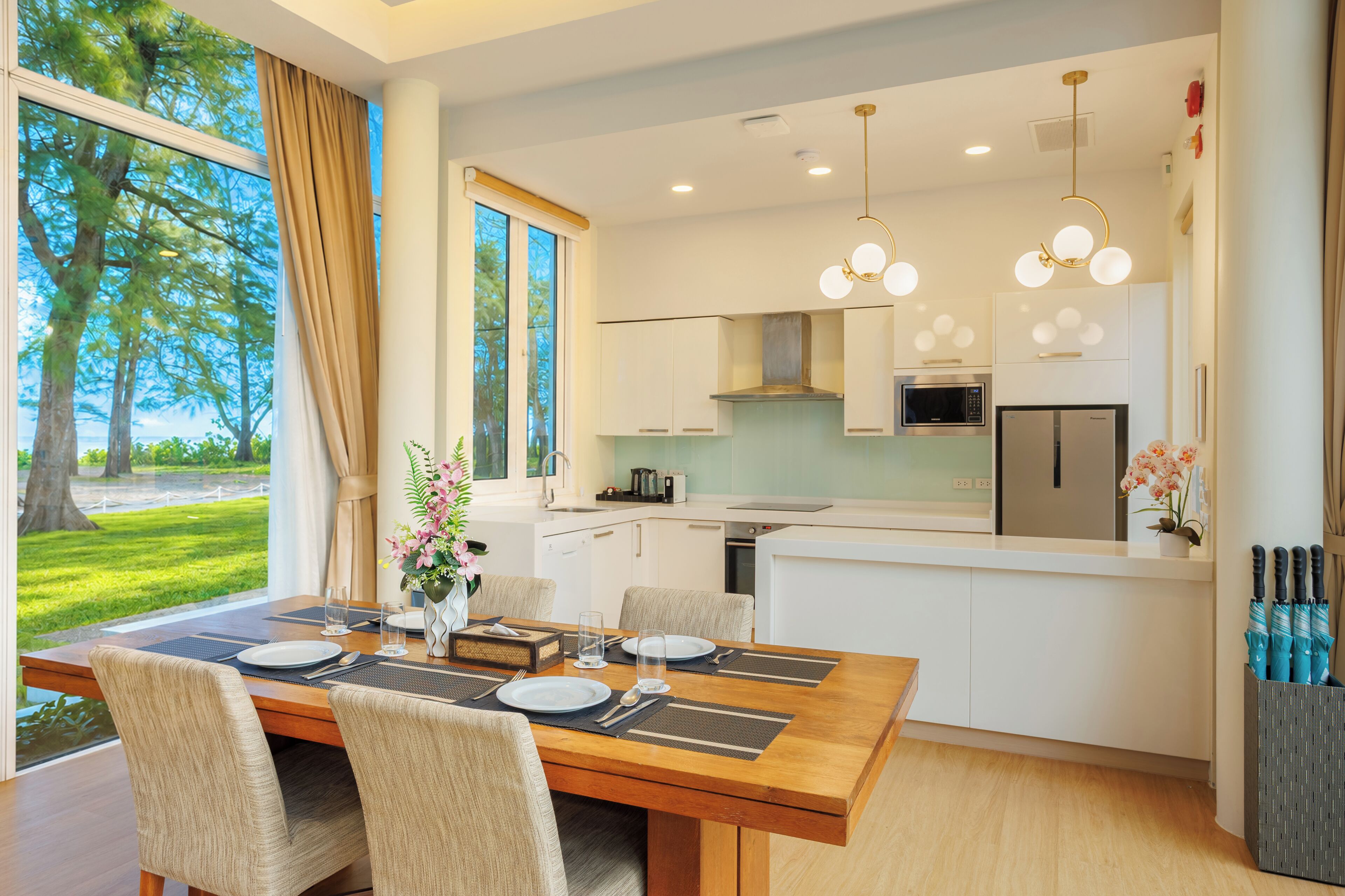 A modern kitchen with white cabinets, a dining area with a wooden table, and a view of a green landscape through large windows.