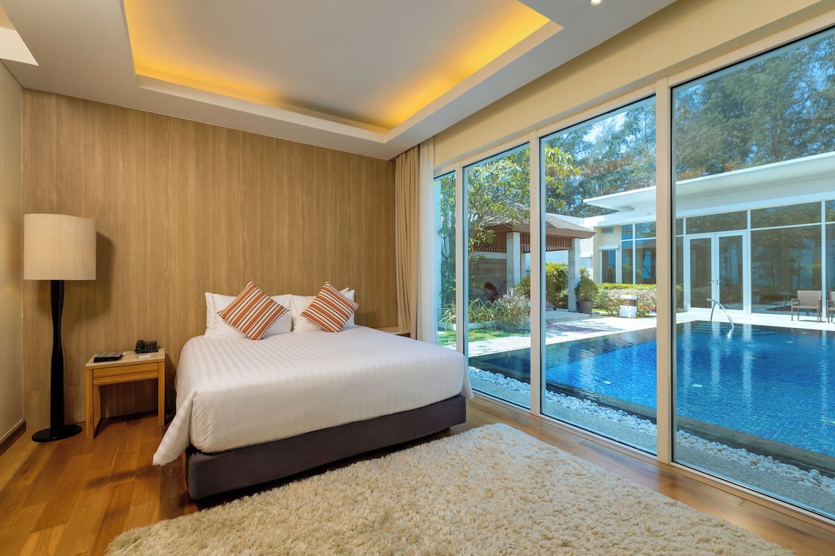 Relax in a spacious room with a large bed, a bedside table, and a lamp, offering a view of the outdoor pool and garden.