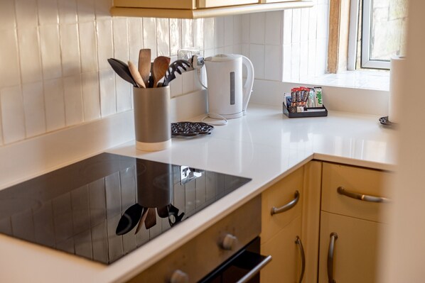 A modern kitchen with a black induction cooktop, a kettle, and a coffee maker. There are wooden utensils in a holder and a coffee pod holder on the counter.