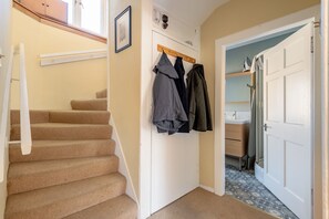 Ein Flur mit beigefarbenem Teppich und einer Treppe, einem Kleiderständer mit mehreren Mänteln, einer weißen Tür und einem Badezimmer mit Fliesenboden.