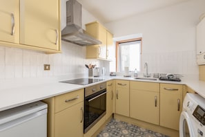 A kitchen with white tiles, yellow cabinets, a stainless steel range hood, a built-in oven, and a window providing natural light. - Market Street (No 97) (St Andrews)