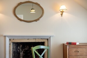 Ein Zimmer mit Kamin, einem grünen Sessel, einer Holzkommode, einem Spiegel und einer Wandlampe.