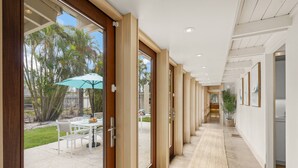 A corridor with glass doors leading to an outdoor patio area with a table and chairs, and a view of palm trees.