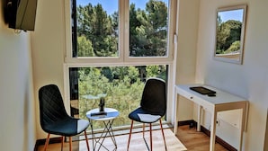 A room with two chairs, a small table, a desk, and a large window offering a view of trees and greenery.