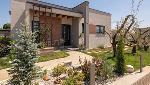 Una casa moderna de dos pisos con un porche cubierto, un pequeño jardín con diversas plantas y un cielo despejado.