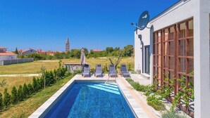 Una piscina con sillones de descanso, un edificio moderno con grandes ventanales y una vista despejada de una torre distante.