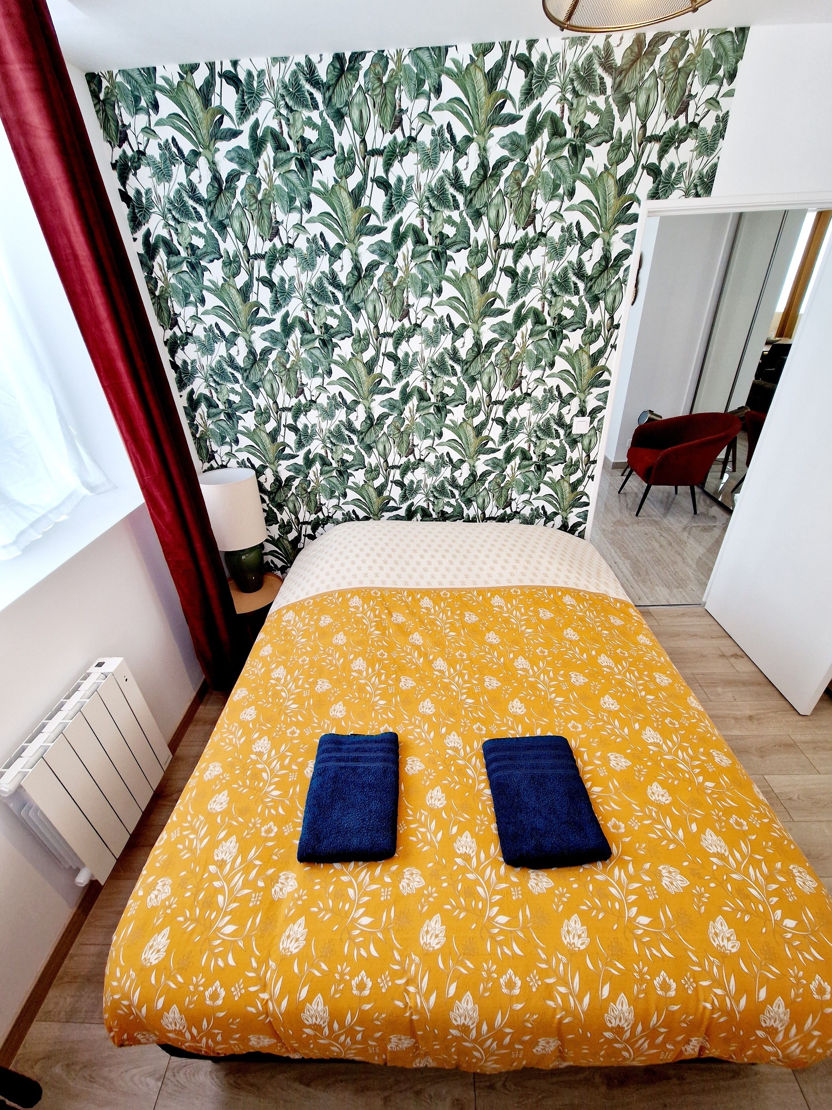 Een slaapkamer met een bed, een rood gordijn, een witte radiator en een behang met een bladmotief.