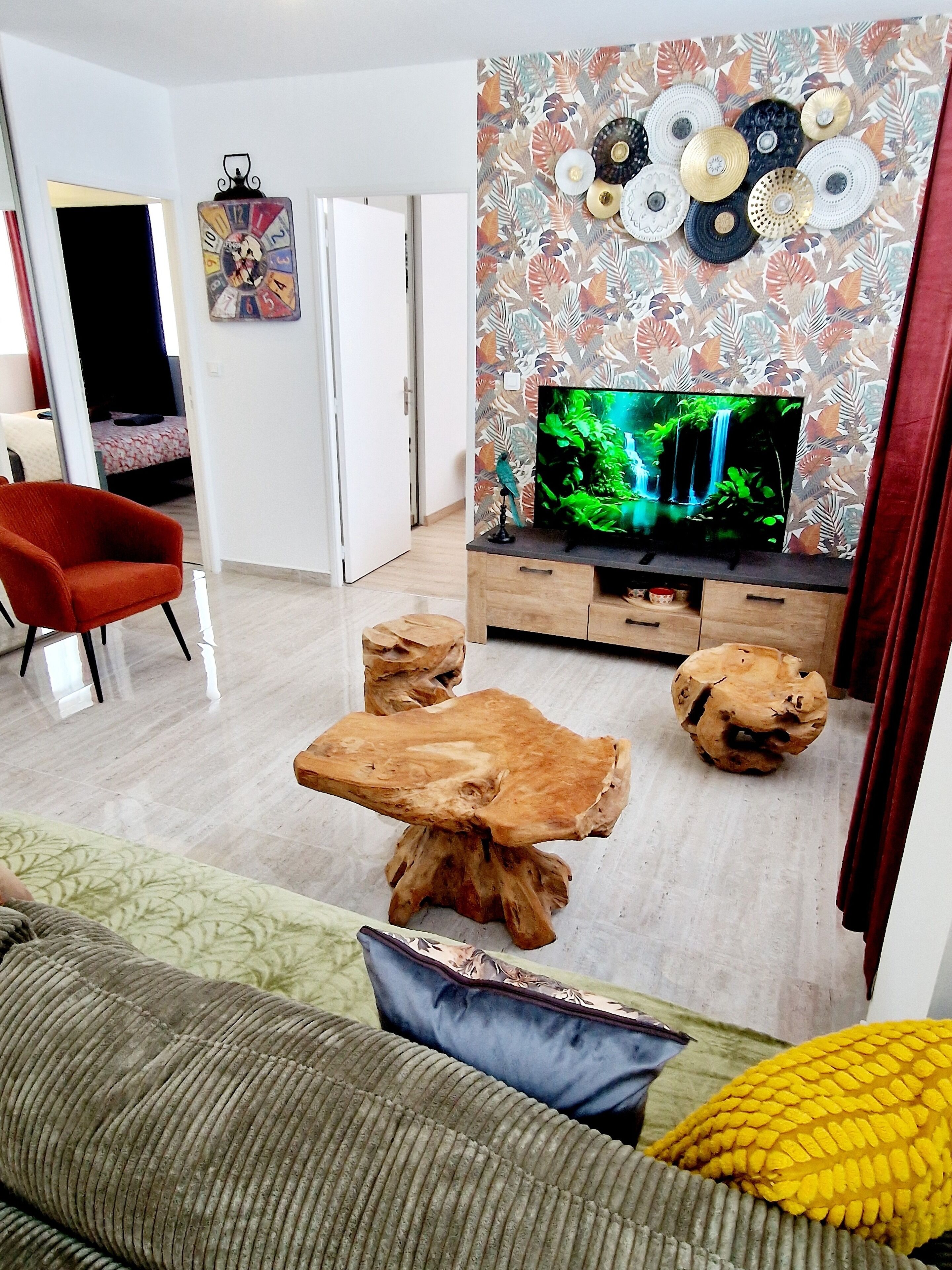 Un salon avec un téléviseur posé sur un meuble en bois, un fauteuil rouge, un canapé et un mur décoratif orné d’un motif floral.