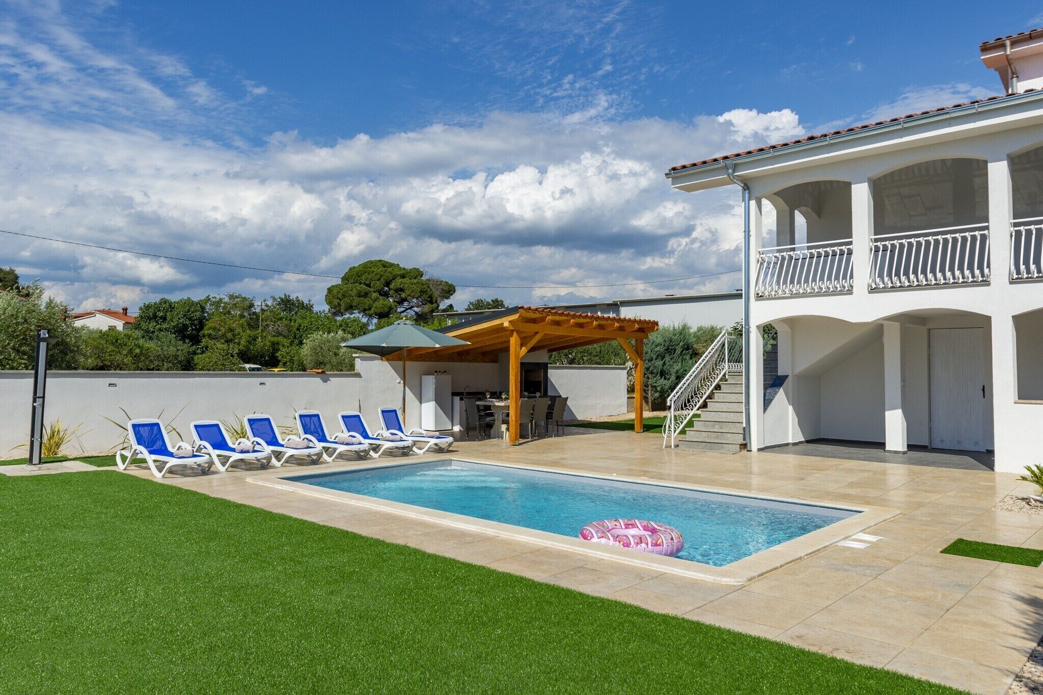 Uma piscina com espreguiçadeiras, um terraço coberto e um edifício de dois andares com varandas.