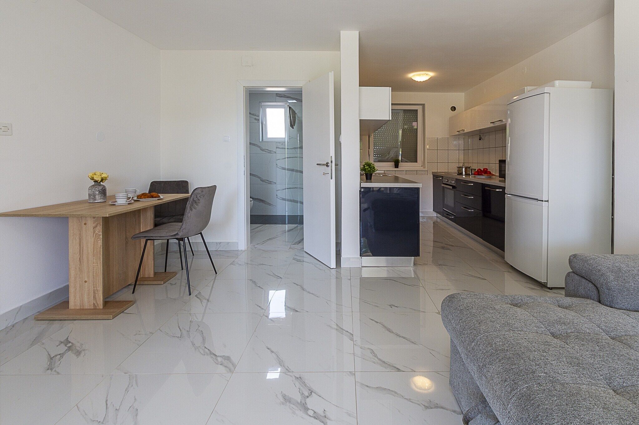 A modern kitchen with a dining area, marble flooring, and a grey sofa.