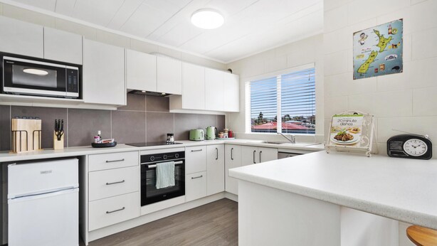 Een moderne keuken met witte kasten, een ingebouwde magnetron, een kookplaat en een spoelbak. Er is een raam met jaloezieën, een kleine radio en een kaart aan de muur.