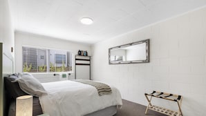 Een slaapkamer met een bed, nachtkastje, lamp, raam met zonwering, radiator en een spiegel aan de muur.