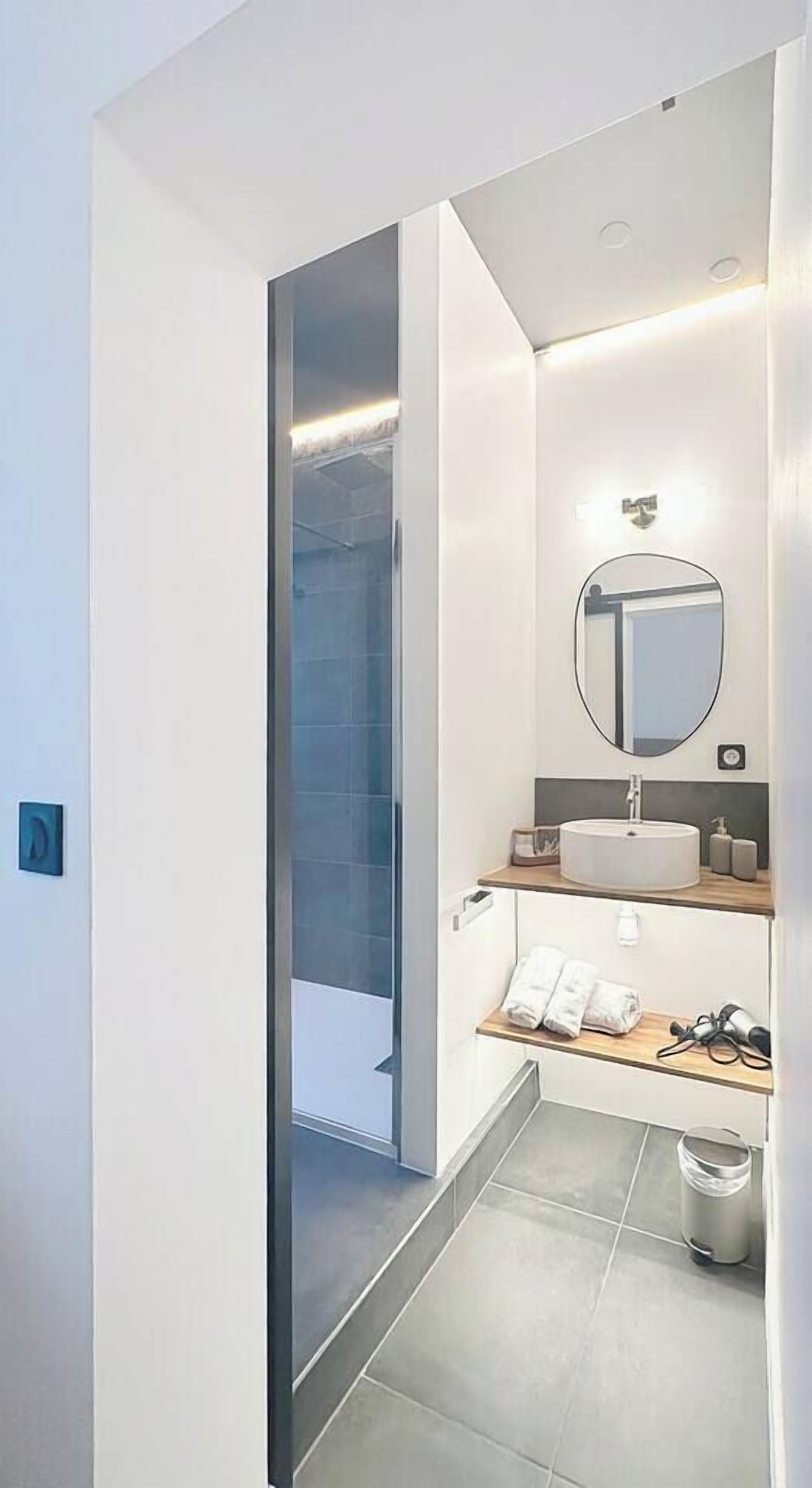A modern bathroom with a glass shower enclosure, a round mirror, a sink with a countertop, and a small shelf with towels and toiletries.