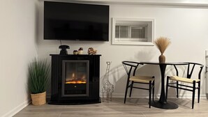 A modern living room with a fireplace, a dining table with chairs, a TV mounted on the wall, and a window with blinds.