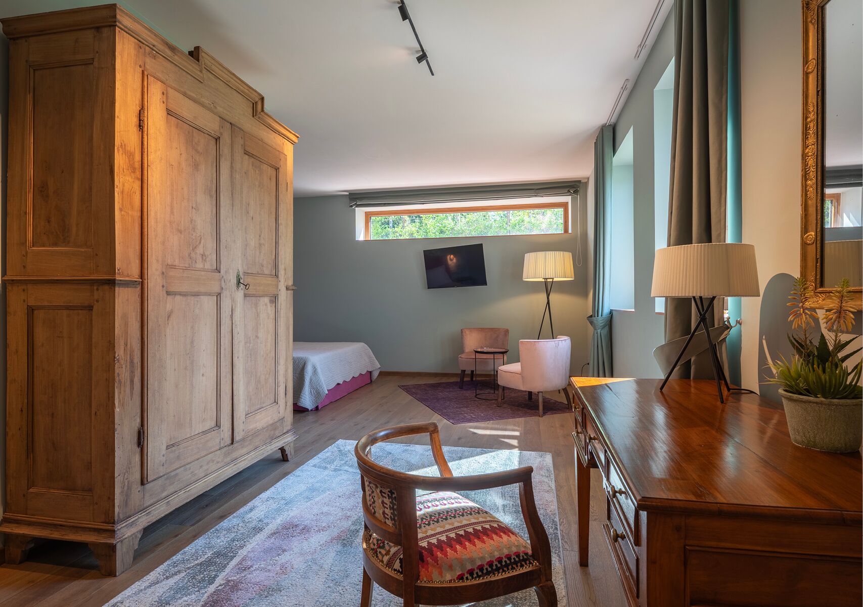 Una habitación con un armario de madera, una cama con colcha rosa, una mesita con lámpara y una silla con cojín estampado.