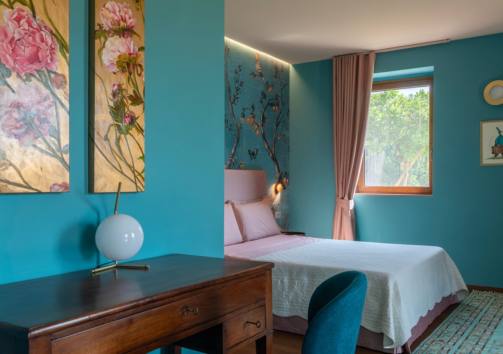 A bedroom with a bed, a desk, and a chair. There are floral paintings on the wall and a window with curtains.