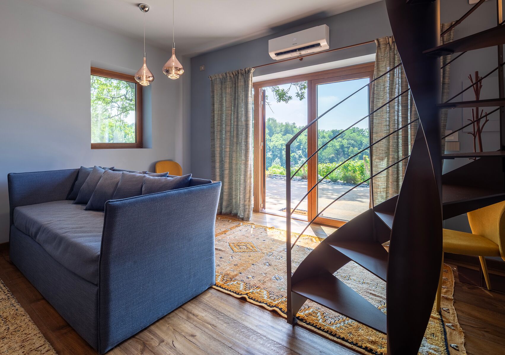 Uma sala de estar moderna com um sofá azul, piso de madeira, um tapete estampado e uma escada com corrimão de vidro que dá para uma varanda.