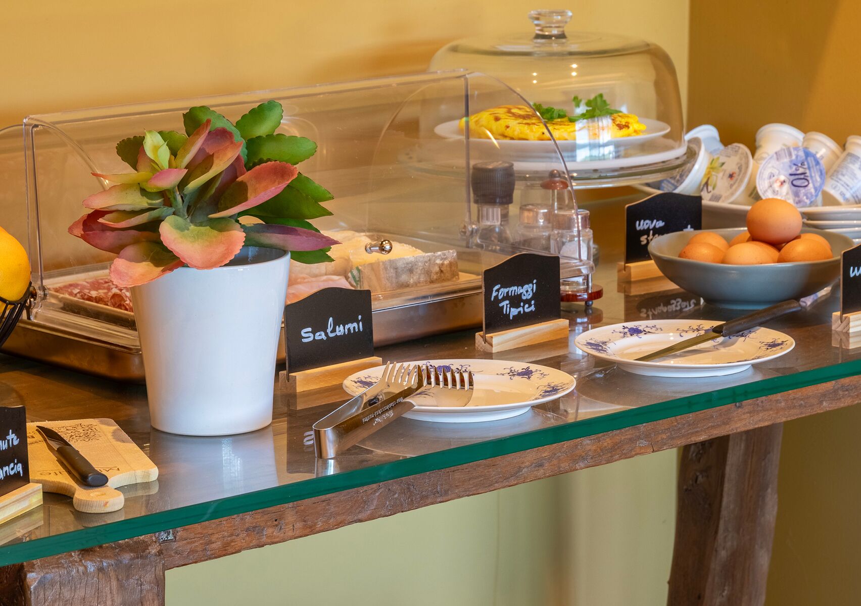 A buffet area with labeled dishes, a plant, and a bowl of oranges.