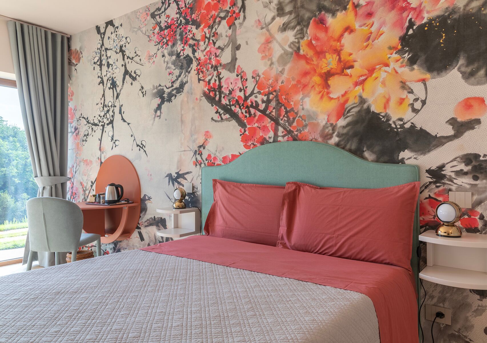 Habitación de hotel con una cama grande, un escritorio y una silla. Hay una ventana con cortinas y una pared con un mural floral.