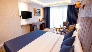 Quarto de hotel com uma cama grande, escrivaninha, cadeira, televisão e janela com cortinas.