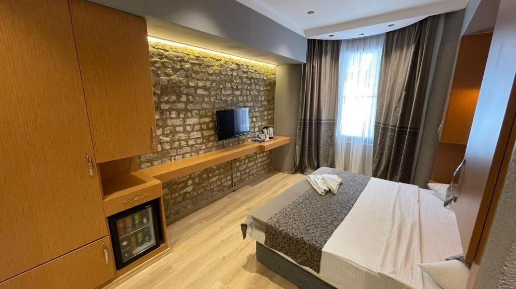 Ein Hotelzimmer mit einem Bett, einem an einer Steinwand montierten Fernseher, einer Minibar und einem Fenster mit Vorhängen.