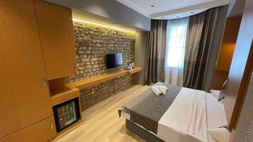 Ein Hotelzimmer mit einem Bett, einem an einer Steinwand montierten Fernseher, einer Minibar und einem Fenster mit Vorhängen.