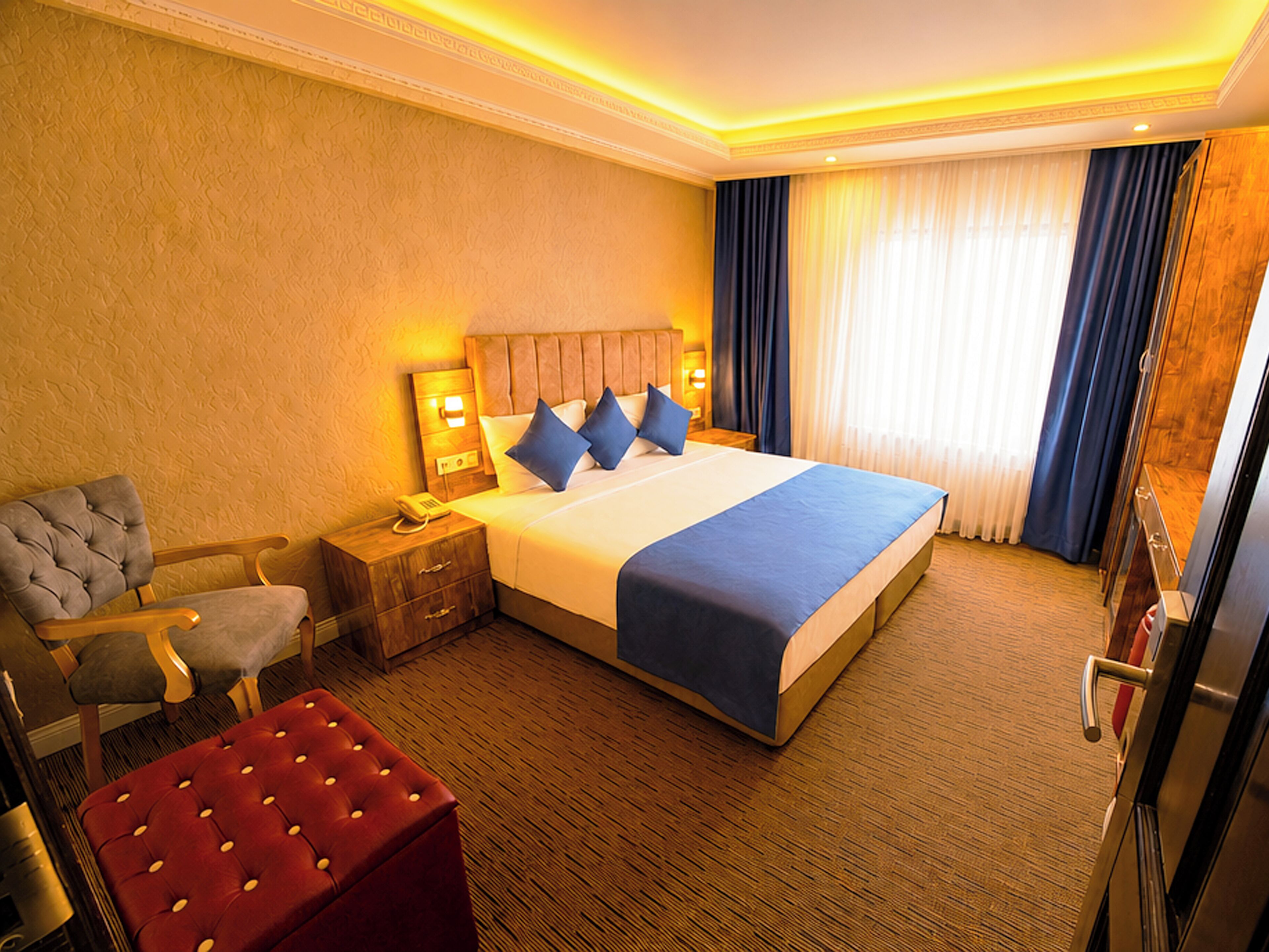 A hotel room with a large bed, a wooden nightstand, a chair, and a red ottoman.