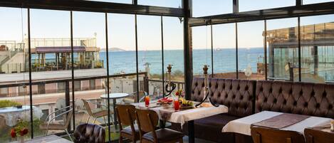 Un restaurante con vistas al mar, con mesas y sillas, una zona de bar y un área de asientos con respaldo alto.