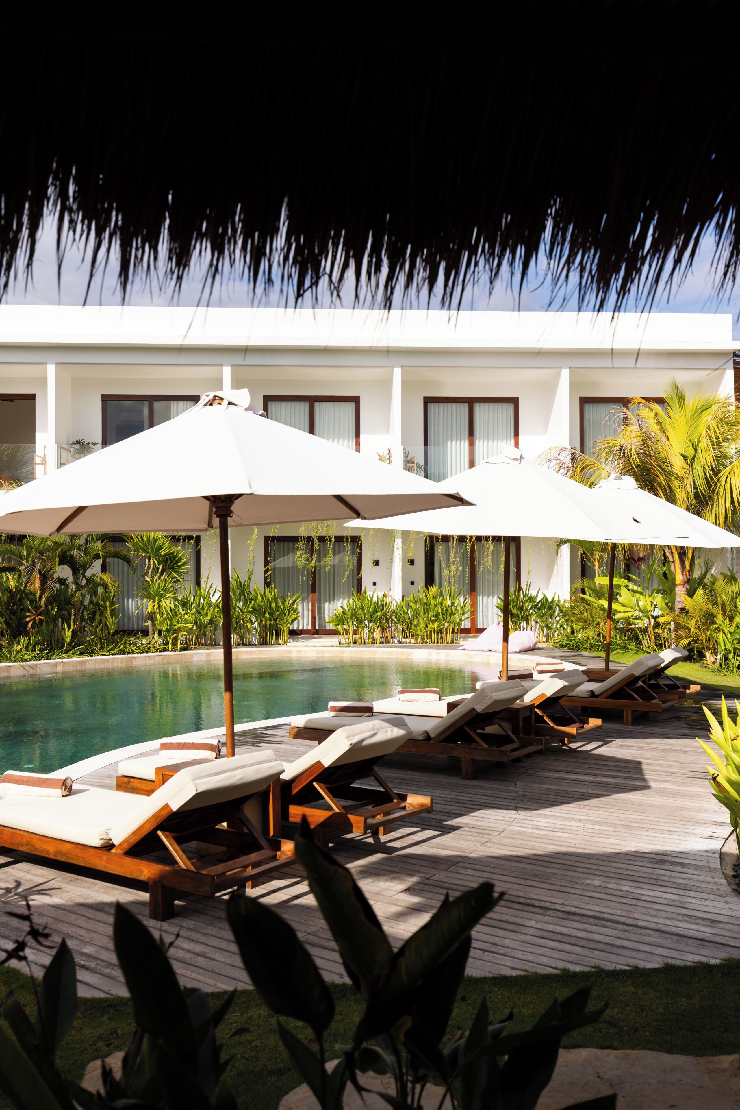Un espace aménagé au bord de la piscine, avec des chaises longues et des parasols, entouré d’une végétation luxuriante et bordé d’un bâtiment doté de grandes fenêtres.