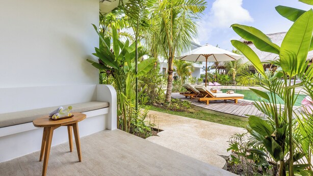 A balcony with a wooden table, a cushioned bench, and a pool area with lounge chairs and umbrellas. - Uryah Hotel Lombok (Selong Belanak)