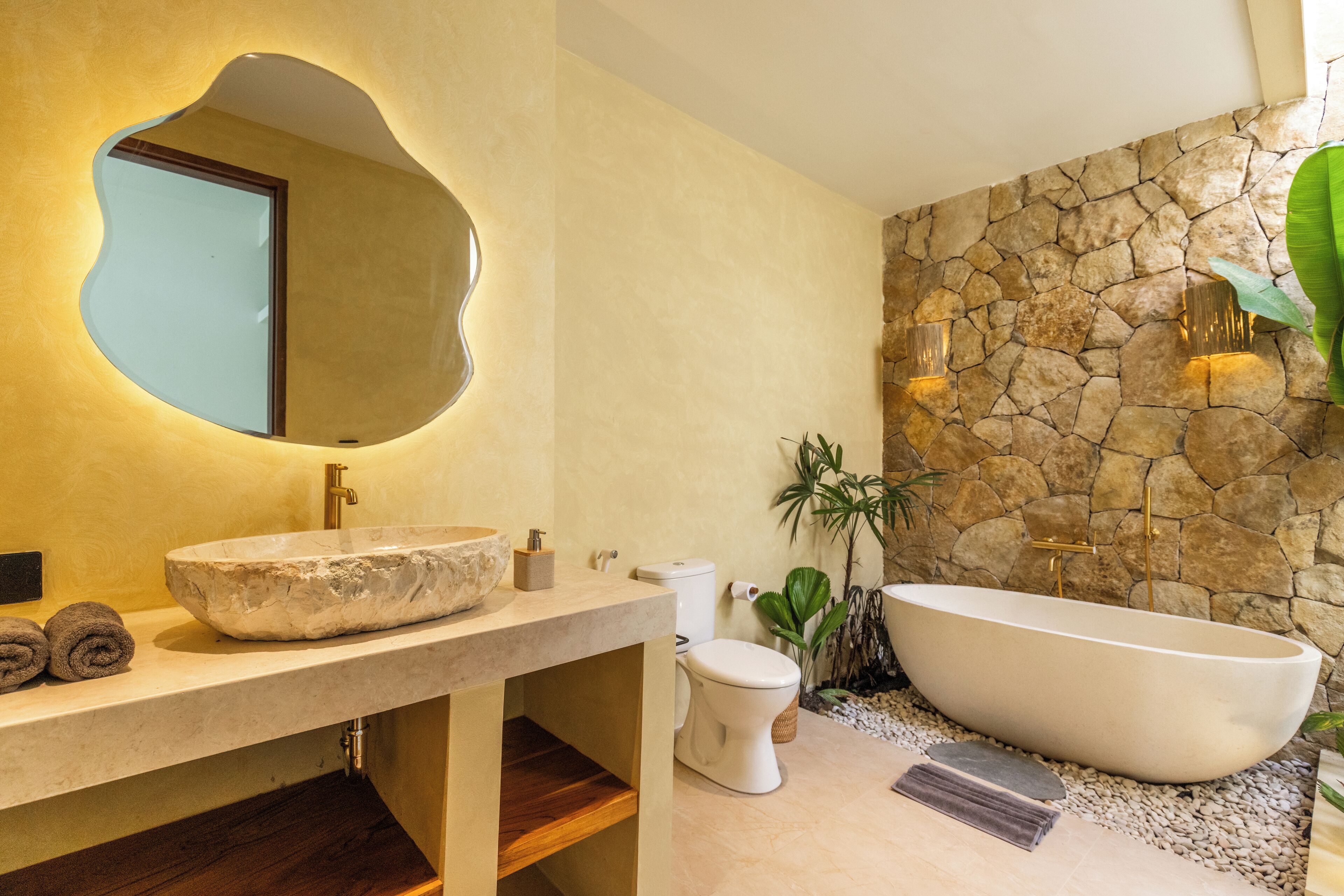 Un baño con pared de piedra, una bañera independiente, un lavabo ovalado grande y un espejo de forma original.