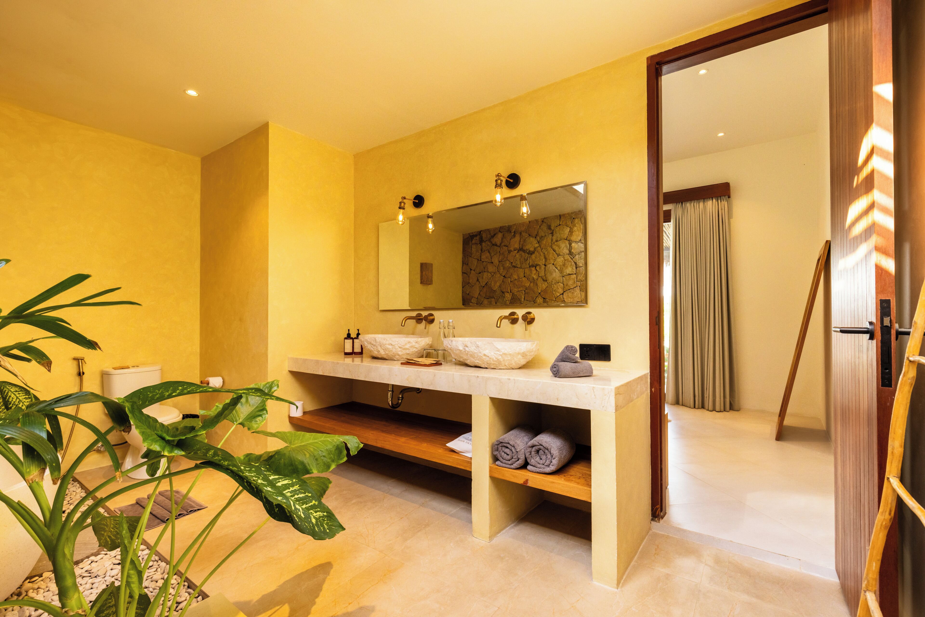A bathroom with a large mirror, two sinks, a wooden shelf with towels, and a potted plant.