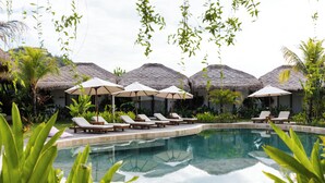 A pool area with lounge chairs and umbrellas, surrounded by thatched-roof structures. - Uryah Hotel Lombok (Selong Belanak)
