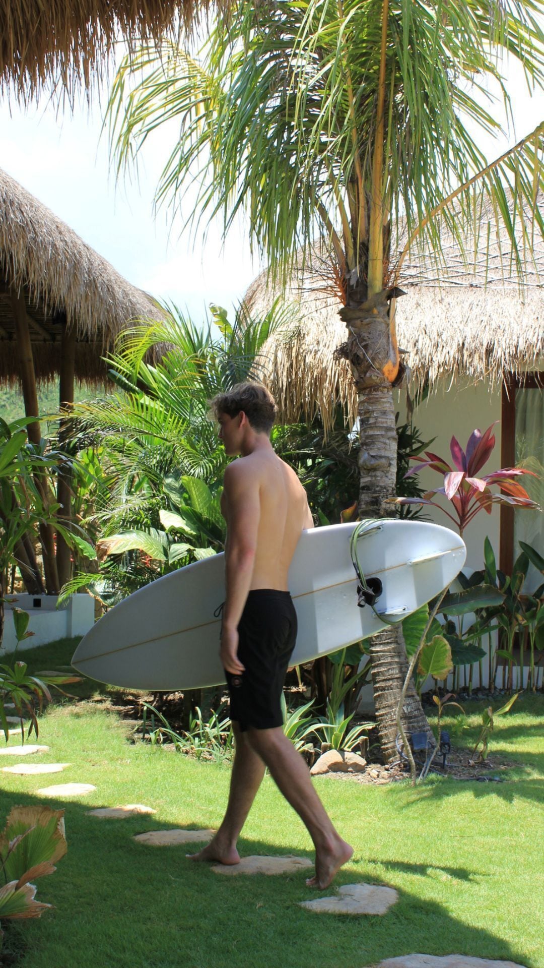 Un homme marchant avec une planche de surf, des palmiers et une structure à toit de chaume.